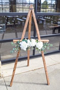 a wooden easel with flowers and greenery in front of a glass building window