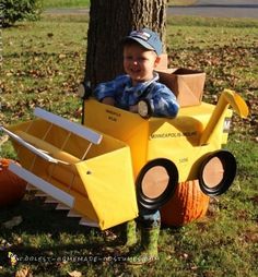 a young boy sitting in a fake construction vehicle costume next to a tree and pumpkins