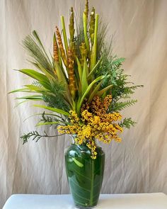 a green vase filled with yellow flowers and greenery on a white tableclothed backdrop