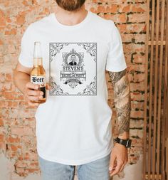 a man with a beard is holding a beer in his hand and wearing a white t - shirt