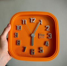 an orange square clock with numbers on the face and hands holding it in front of a gray wall