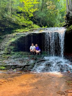 two people sitting on rocks in front of a waterfall with water cascading over them