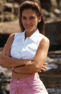 a woman with her arms crossed standing in front of some rocks and water wearing pink shorts