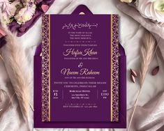 an ornate purple and gold wedding card on top of a white table cloth with flowers