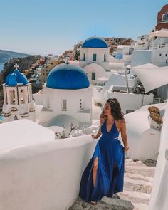 a woman in a blue dress is standing on steps near the water and buildings that look like whitewashed houses