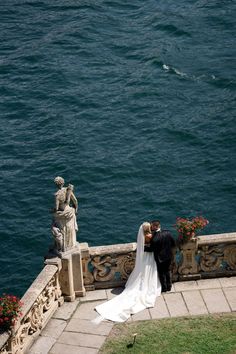 a bride and groom standing on the edge of a stone wall next to water with statues