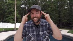 a man in plaid shirt and hat holding his hands up to his ears with trees in the background