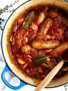 sausages and beans are cooked in a sauce with sage leaves on the side, next to a wooden spoon