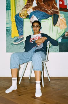 a man sitting in a chair next to a painting on the wall and wearing glasses