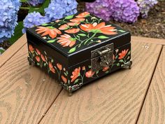 a black box with flowers painted on it sitting on a wooden table next to purple and blue flowers