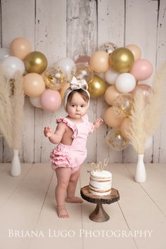 a baby girl standing in front of a cake with balloons behind her and posing for the camera