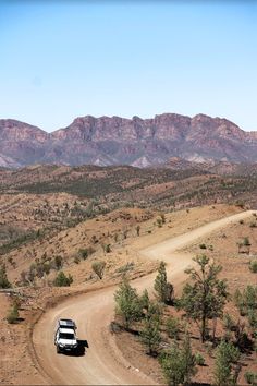 a truck driving down a dirt road in front of some mountain range with trees and bushes