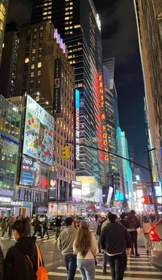 many people are walking on the street at night in new york city, with skyscrapers lit up