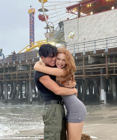 a man and woman hugging each other on the beach near an amusement park ferris wheel