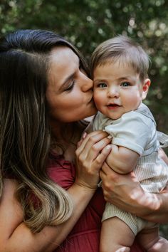 a woman holding a baby and kissing it's face with trees in the background