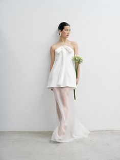 a woman standing in front of a white wall wearing a dress and holding a flower