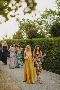 a group of women walking down a dirt road next to bushes and trees with one woman in a yellow dress