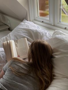 a woman laying in bed with an open book on her lap and looking out the window