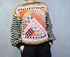 a woman standing in front of a gray wall wearing a colorful sweater and black pants