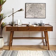 a wooden desk topped with a lamp next to a potted plant and a framed map