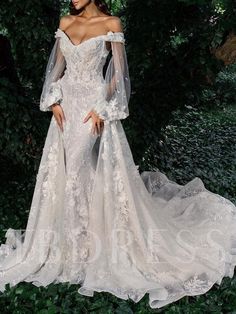 a woman in a white wedding dress standing next to some green bushes and trees with her hands on her hips