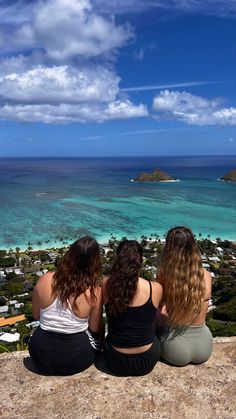 three women sitting on the edge of a cliff looking out at an island and ocean