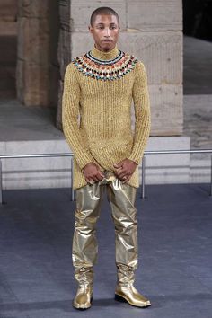 a man standing in front of a building wearing gold pants and a sweater with beads on it