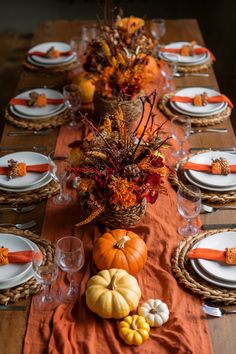 a wooden table topped with lots of white plates and orange napkins next to a basket filled with pumpkins