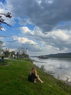 a woman is sitting on the grass next to the water and looking at something in the distance