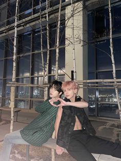 two young men sitting on a bench in front of a building at night with the city lights behind them