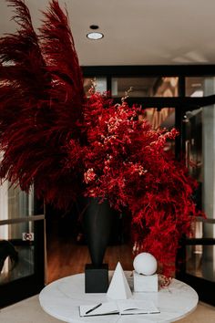 a white table topped with a black vase filled with red flowers next to a tall plant