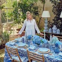 a woman standing in front of a table with blue and white dishes on it, surrounded by potted plants