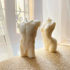 two white candles sitting on top of a table next to a window with curtains in the background