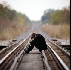 a man sitting on train tracks with his head down and hands in his pockets, looking at the ground