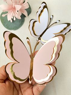 a hand holding a pink and gold butterfly on top of a white table with flowers in the background