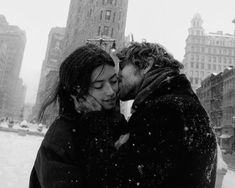 a man and woman kissing in the snow on a city street with tall buildings behind them