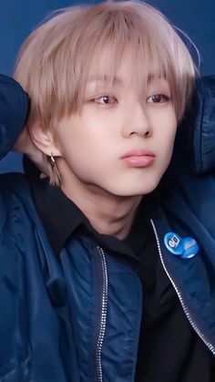 a young man with blonde hair wearing a blue jacket and earrings, looking at the camera