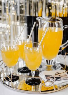 three glasses filled with orange juice on a silver serving tray next to black and white bottles