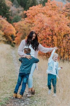 a woman and two children walking down a dirt road in the woods with fall foliage behind them