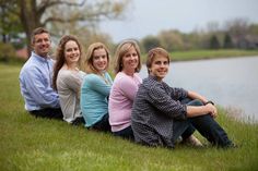 a group of people that are sitting in the grass by some water with trees behind them