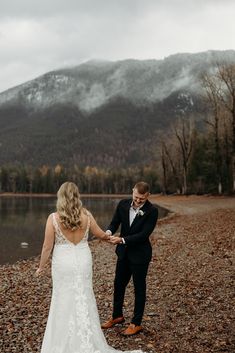 a bride and groom holding hands on the shore of a lake with mountains in the background