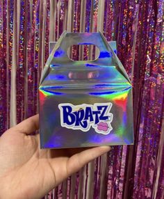 someone holding up a box with the word bratz on it in front of purple and pink tinsel