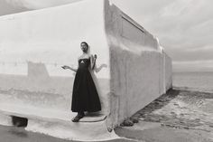 a woman in a black dress standing next to a white wall by the ocean with her hand on her hip