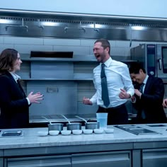 two men and one woman are laughing in the kitchen while another man is talking to them