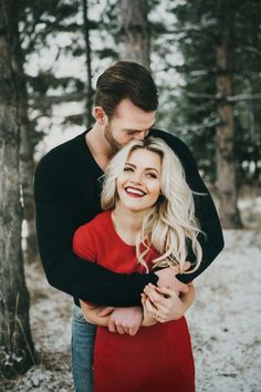 a man and woman hugging in the woods with snow on the ground, trees behind them