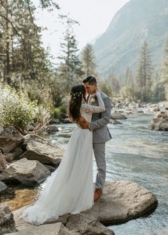 a bride and groom standing on rocks by the river in their wedding attire, embracing each other