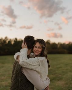 a woman is hugging her friend in the park at sunset with clouds overhead and trees behind her