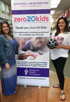 two women standing in front of a sign holding a soccer ball and smiling at the camera