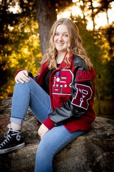 a young woman sitting on top of a rock in front of some trees wearing a red and black jacket