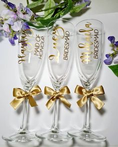 three wine glasses with gold bows and names on them, sitting next to purple flowers